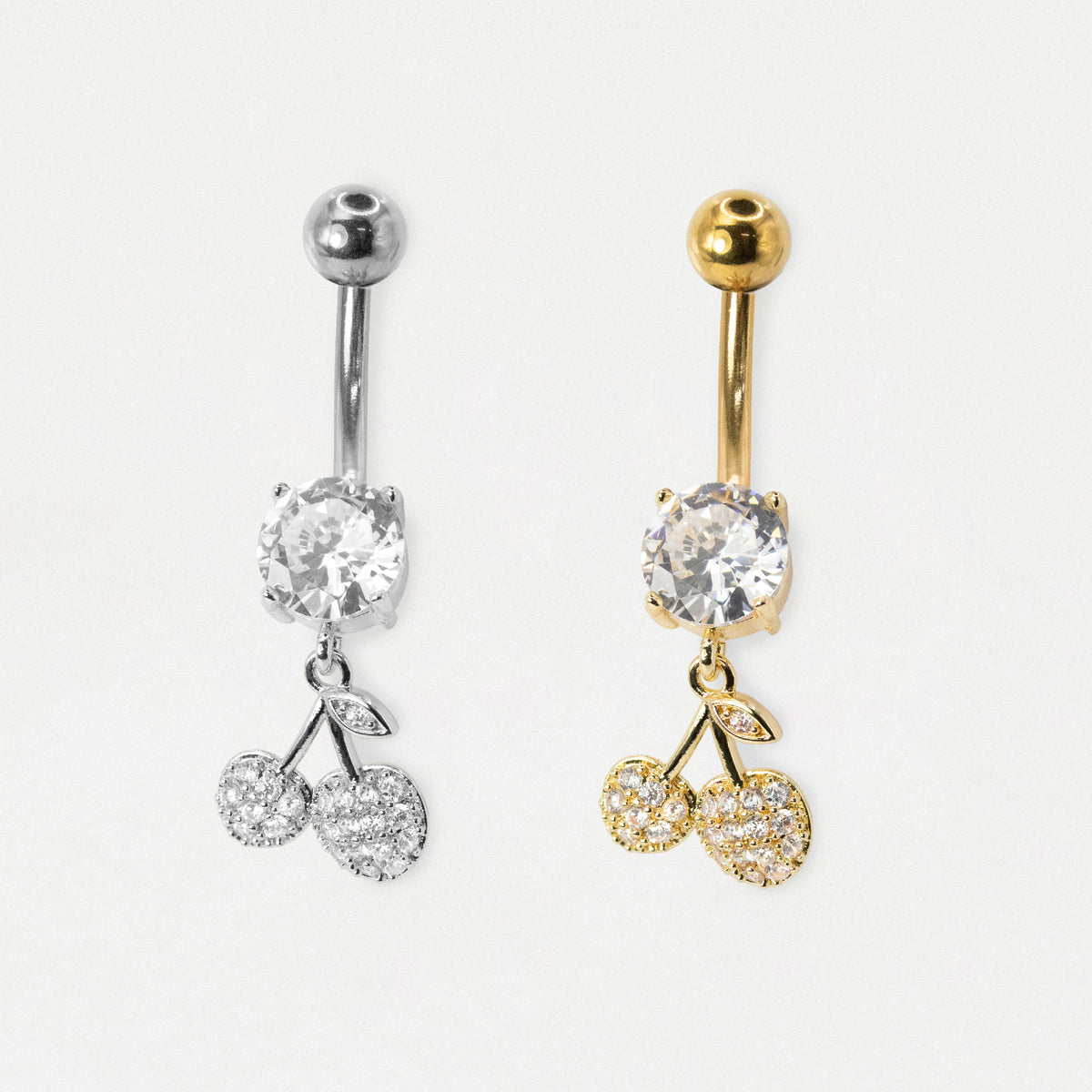 Two cherry-shaped belly button rings shown side by side on a light gray background. The left ring is silver-toned with a clear round stone and two clear stone cherries.