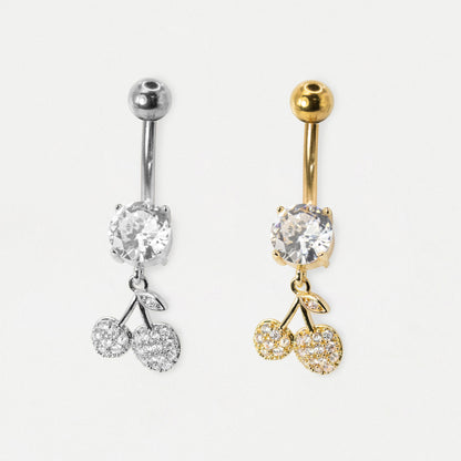 Two cherry-shaped belly button rings shown side by side on a light gray background. The left ring is silver-toned with a clear round stone and two clear stone cherries.