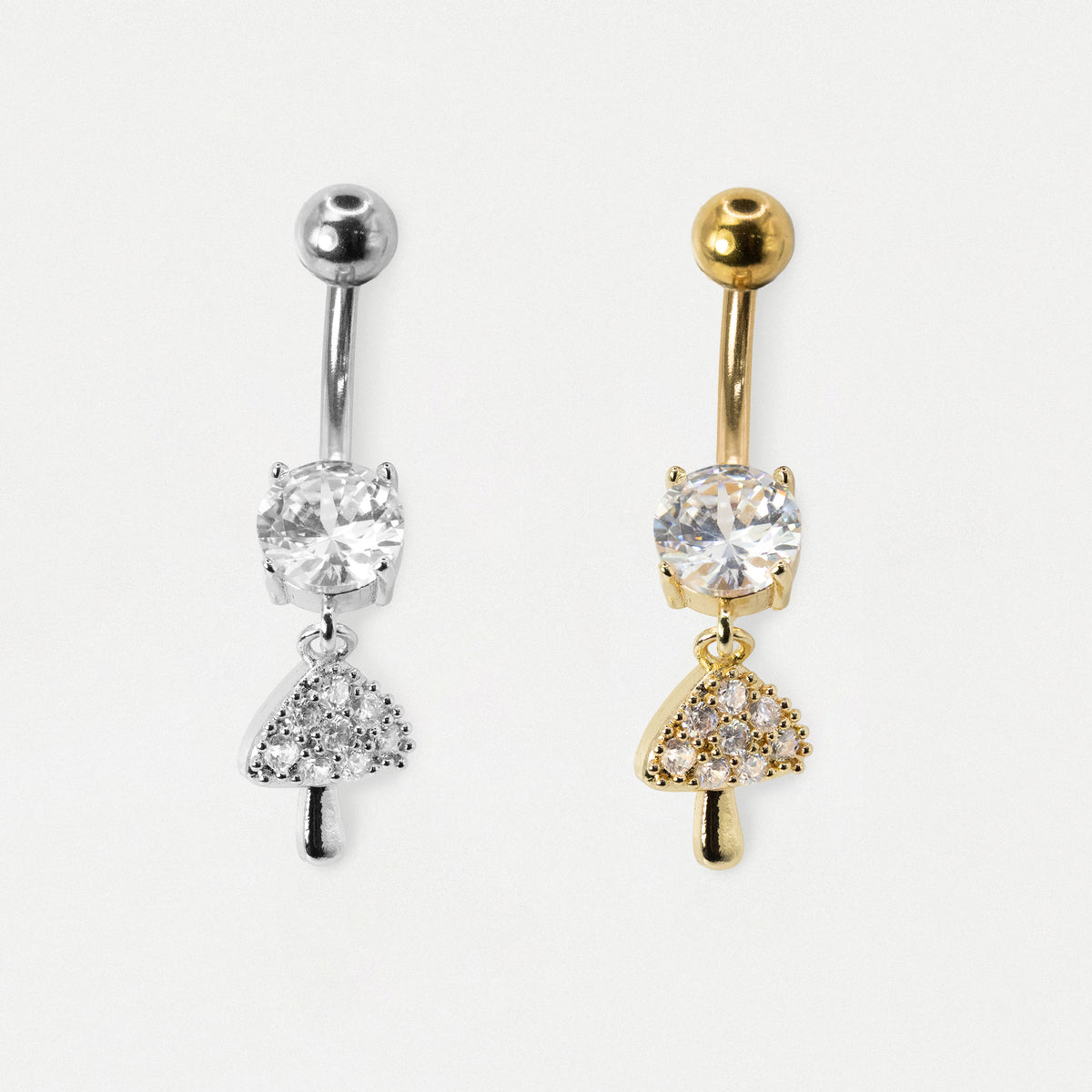 Two belly button rings with mushroom-shaped dangling charms, one in silver and one in gold, both featuring clear gemstones, shown on a light gray background.