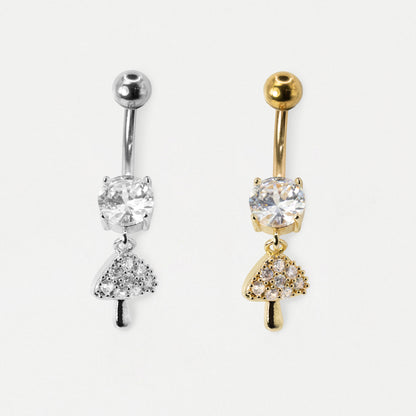 Two belly button rings with mushroom-shaped dangling charms, one in silver and one in gold, both featuring clear gemstones, shown on a light gray background.