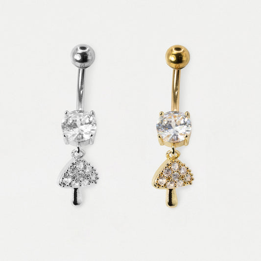 Two belly button rings with mushroom-shaped dangling charms, one in silver and one in gold, both featuring clear gemstones, shown on a light gray background.