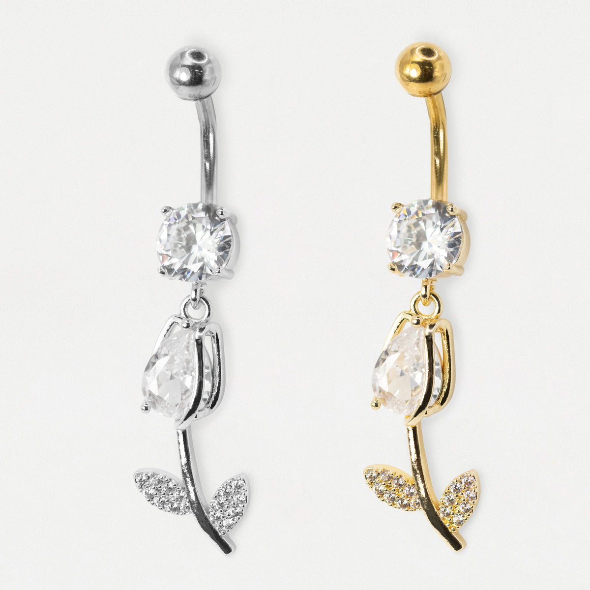 Two rose-shaped belly button rings shown side by side on a plain light background. One is silver-toned and the other is gold-toned, both featuring a clear faceted gemstone, a teardrop-shaped clear gemstone, and leaf details paved with small clear stones.