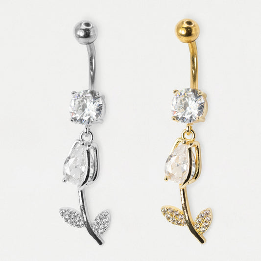 Two rose-shaped belly button rings shown side by side on a plain light background. One is silver-toned and the other is gold-toned, both featuring a clear faceted gemstone, a teardrop-shaped clear gemstone, and leaf details paved with small clear stones.