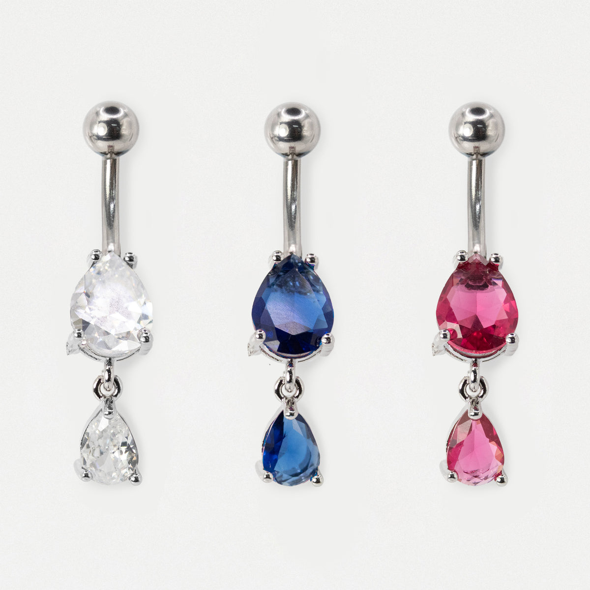 Three steel belly button rings with teardrop-shaped gemstones are displayed side by side on a light gray background. The left belly bar has a clear faceted stone, the middle ring has a deep blue faceted stone, and the right ring has a pink faceted stone.