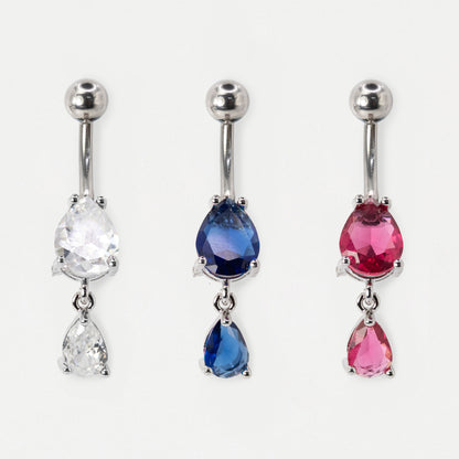 Three steel belly button rings with teardrop-shaped gemstones are displayed side by side on a light gray background. The left belly bar has a clear faceted stone, the middle ring has a deep blue faceted stone, and the right ring has a pink faceted stone.